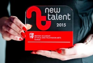 A person holds a transparent acrylic recognition award from the british academy of film and television arts scotland for new talent 2015, prominently featuring a red design.