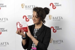 A young woman proudly holding a new talent recognition award from bafta scotland, smiling as she looks at the trophy. she is at an event backdrop with logos.