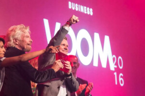 Sir richard branson celebrating with an entrepreneur on stage at the voom 2016 business event, under a pink-lit backdrop with the event's name displayed.
